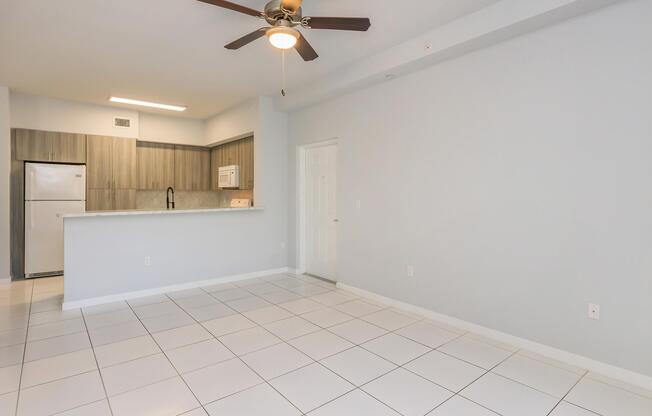 Bright and spacious interior view of a modern apartment with light gray walls and large white tile flooring. A ceiling fan hangs above the room. The open kitchen features wooden cabinets and a refrigerator visible in the corner. The space is empty, creating a clean and inviting atmosphere.