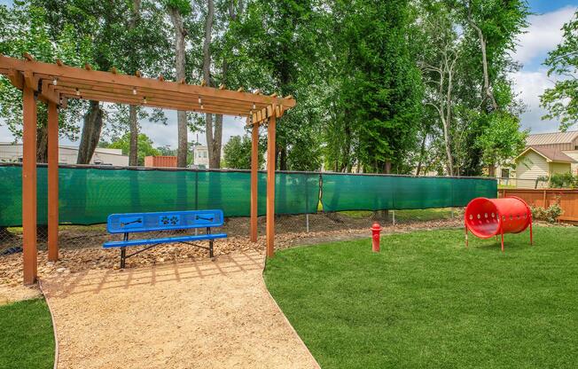 A pet-friendly park area featuring a blue bench under a wooden pergola, a red dog tunnel, and a fire hydrant on a grassy lawn. The space is enclosed by a green fence and surrounded by trees, creating a welcoming outdoor environment for dogs and their owners.