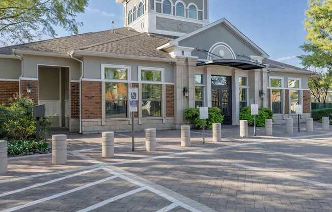 A leasing office exterior here at Landing at Mansfield with brick and stone facade, arched upper windows, covered entry, glass doors, short concrete bollards, and a paved walkway in bright daylight.