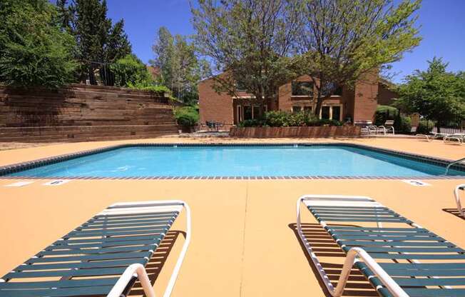 A sunny outdoor swimming pool here at Eagle Ridge showcasing a large rectangular pool with clear blue water, a wide concrete pool deck, two striped lounge chairs in the foreground, additional seating around the perimeter, landscaped trees and greenery, and a brick clubhouse building in the background beneath a clear blue sky.