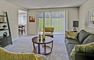A spacious living room here at Andorra showcasing a gray sofa with patterned accent pillows, a round wooden coffee table with decorative vases, a coordinating accent chair, and soft carpeted flooring, with vertical blinds covering a sliding glass door that opens to a grassy outdoor view, while open shelving, wall art, and a table lamp add warmth and function.