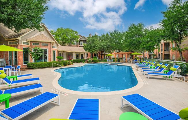 A clear blue swimming pool surrounded by lounge chairs in bright blue and green. Lush greenery and trees provide shade around the pool area, with a few umbrellas nearby. Residential buildings are visible in the background under a partly cloudy sky.