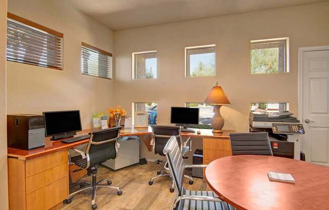 A bright business center here at Dobson Towne Center featuring a long wood workstation with multiple desktop computers, black swivel chairs, and a convenient printer station, complemented by a round meeting table, warm table lamp, and tall windows that bring in natural light to create a comfortable and productive workspace for residents.