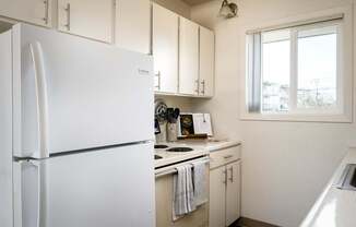 A white refrigerator stands in a kitchen with white cabinets and a window.