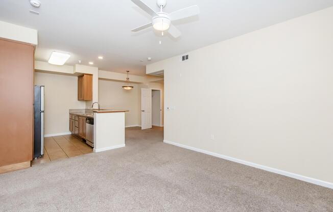 A spacious interior of a modern apartment featuring a kitchen with wooden cabinets, stainless steel appliances, and a kitchen island. The living area has light-colored walls, a ceiling fan, and soft carpeting. A doorway leads to another room, suggesting an open layout.