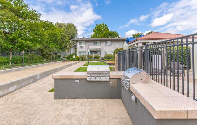 Outdoor grilling area featuring two stainless steel barbecue grills on a tiled counter, surrounded by green landscaping. In the background, there are residential buildings and a gravel pathway lined with trees under a partly cloudy sky.