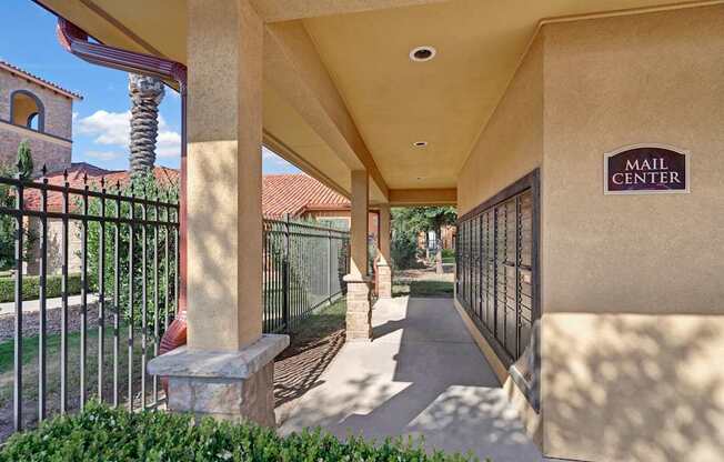 A convenient mail center walkway here at Tuscany at Faudree featuring covered stucco columns, a row of secure metal mailboxes, and a shaded concrete path. Wrought iron fencing, landscaped greenery, and Mediterranean-style architecture with tile roofs create a clean, accessible, and welcoming community setting.