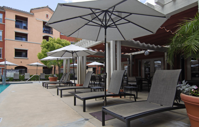 A patio with chairs and umbrellas is surrounded by a pool and buildings.