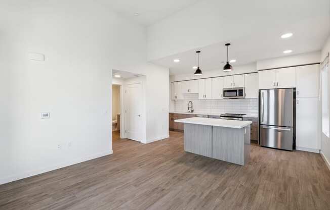 an empty living room with a kitchen with a stainless steel refrigerator
