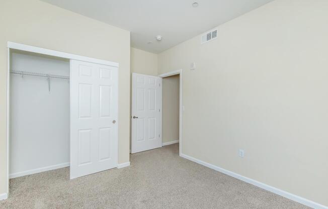 A light-colored room with beige walls and carpet flooring. There are two closed white doors, one of which leads to a closet. The room is well-lit and appears clean and empty, suitable for various purposes.