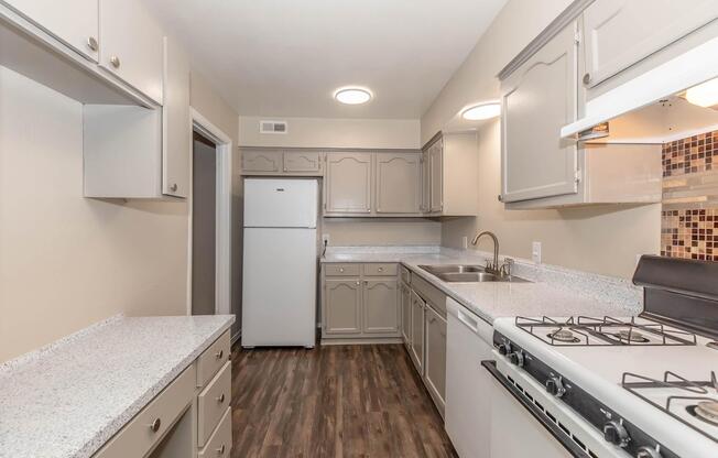 A modern kitchen featuring light gray cabinetry, a white refrigerator, and a stove with a grill. The countertops are light-colored, and there's a tiled backsplash. The floor is a dark wood finish, creating a contemporary and clean aesthetic with ample storage space.