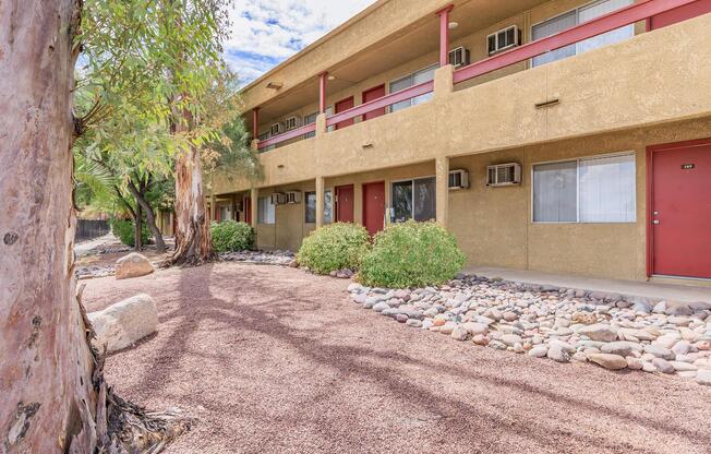 Ground-level view of a building with a tan exterior featuring red doors. Surrounding the building are landscaped areas with shrubs and large rocks, set against a backdrop of trees. The sky is partly cloudy, and there are air conditioning units under the windows.