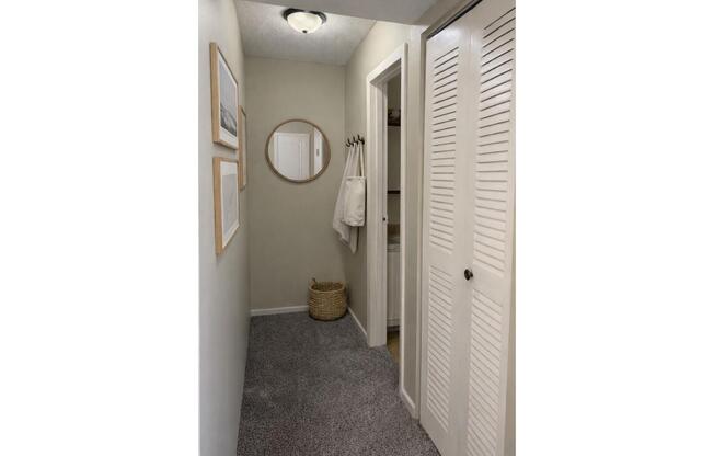 A narrow hallway featuring neutral wall colors, a round mirror, two framed artworks, a closet with louvered doors, and a beige woven basket on the floor. A towel hangs on a hook near the closet, and the space is illuminated by a ceiling light. The carpet is light gray.