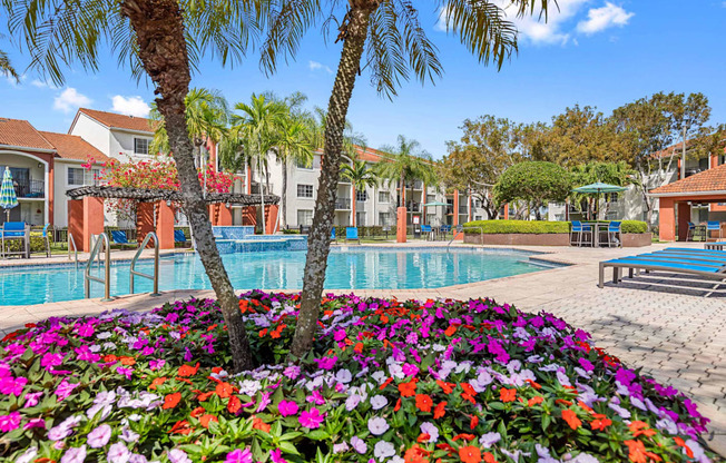 Alternate view of community swimming pool at Sunset Gardens. Hot tub is located near covered lounge area. Lounge seating is located throughout pool deck as well as picnic tables