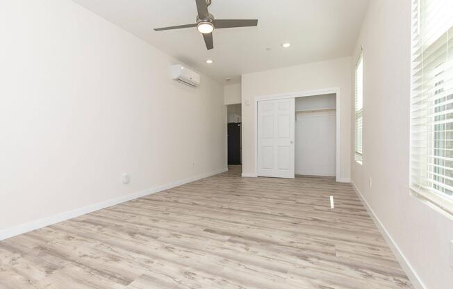 A bright, empty room featuring light-colored walls and a modern ceiling fan. The floor is covered in light wood-like laminate. There is a door leading to a closet on the left side and a window on the right side, allowing natural light to enter. An air conditioning unit is visible on the wall.