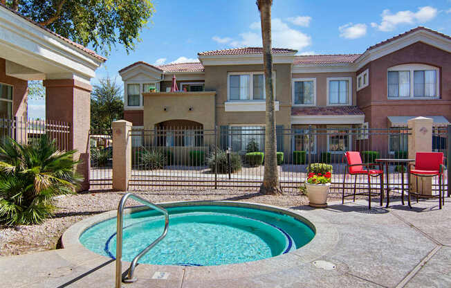 A relaxing spa area here at Dobson Towne Center featuring a circular heated spa with built-in steps and surrounding tiled deck, set beside red patio chairs and a small table for casual outdoor seating. A decorative planter with vibrant flowers, palm trees, and gated courtyard views create a peaceful setting perfect for unwinding under sunny Arizona skies.
