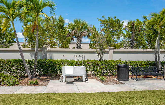 A white bench sits in a park with palm trees and a trash can.