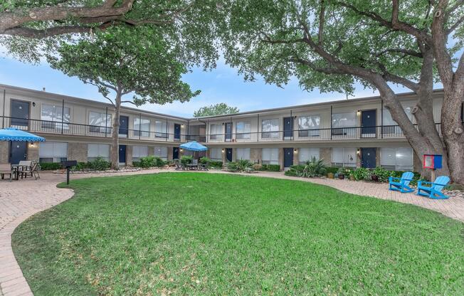 A view of a courtyard at a multi-unit building, featuring green grass, several trees, and outdoor seating with blue chairs. In the center, there is a small patio area with an umbrella and tables. The building has multiple levels with balconies and is surrounded by landscaping.