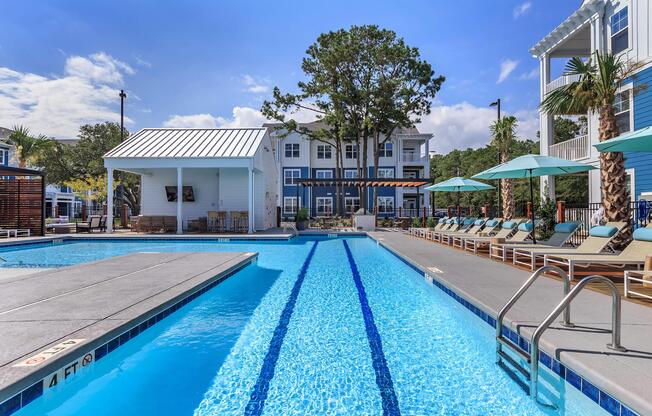 A large outdoor swimming pool with clear blue water, lined with lounge chairs and umbrellas. In the background, there are modern apartment buildings surrounded by palm trees and a sunny sky. A shaded cabana area is visible to the left, enhancing the resort-like atmosphere.