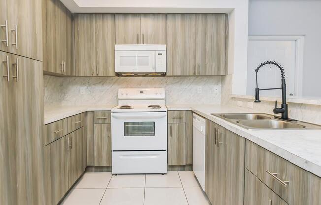 A modern kitchen featuring light wood cabinetry, a white stove and microwave, a sleek black faucet, and a stainless steel sink. The countertops are marble, and the flooring is tiled, creating a clean and contemporary look. The space is well-lit, enhancing the overall ambiance.