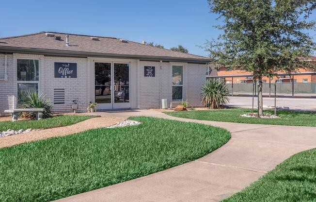 A well-maintained pathway leads to a brick office building with large windows, surrounded by green grass and decorative landscaping. The office door displays a sign that reads "Office." Trees and shrubs enhance the outdoor aesthetic, while the surrounding area features a fenced space in the background.