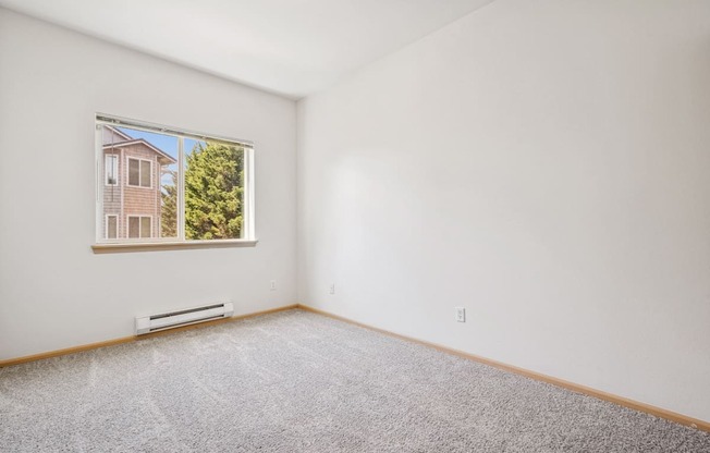 A bedroom with a carpeted floor and a window showing apartments and trees outside at The Madison Apartments in Olympia, WA