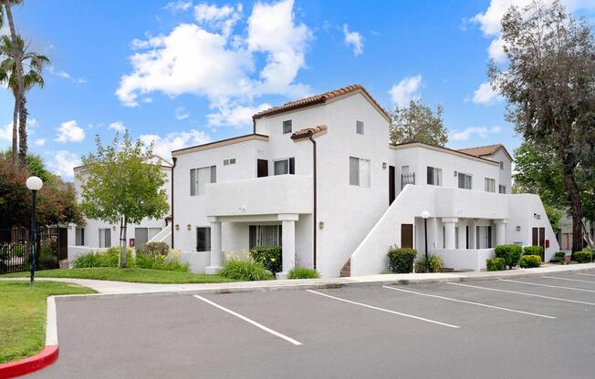 A modern, two-story residential building with white exterior and brown tile roofs, surrounded by green landscaping and palm trees. The parking area has several empty spaces, and the sky is partly cloudy. The entrance features a small staircase leading to balconies.