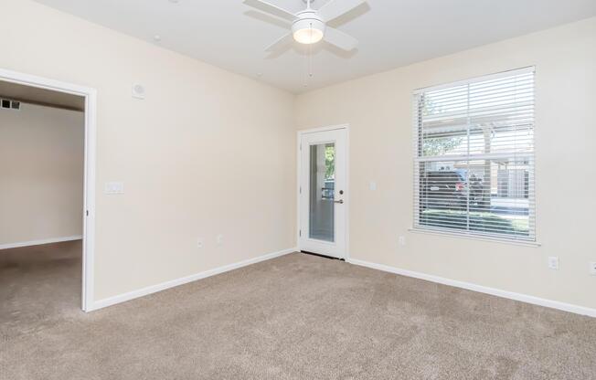 A spacious, light-filled room with beige walls and carpeted flooring. It features a ceiling fan, a large window with blinds, and a glass door leading outside. The room is currently empty, providing a blank canvas for decoration.