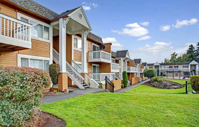 A charming exterior here at Whispering Cedars with well-kept green lawns, landscaped shrubs, and inviting apartment buildings featuring private balconies, exterior staircases, and a peaceful community setting under a bright open sky.