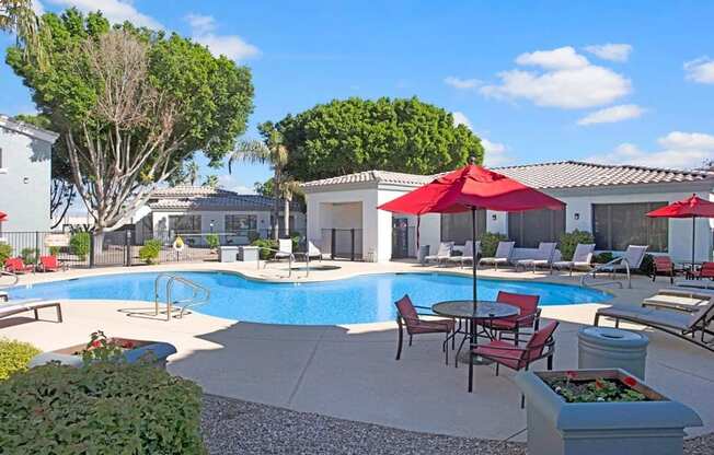 A bright resort-style pool courtyard here at Pala Mesa featuring a sparkling swimming pool surrounded by spacious sundecks, cushioned lounge chairs, and shaded tables with bold red umbrellas. Lush green trees and well-kept landscaping frame the space, while nearby buildings create a private, inviting setting perfect for relaxing or gathering with friends.