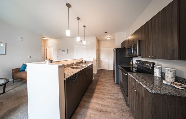 Entrence into kicten, dark brown cabinets with light grey granite countertops, island to the left, stainless steel black appliances, white walls.