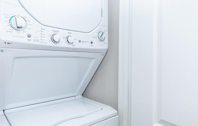 A stacked washer and dryer unit in a laundry room, with control knobs for different settings. The appliance is white and positioned next to a closed door. The overall setting is clean and modern.