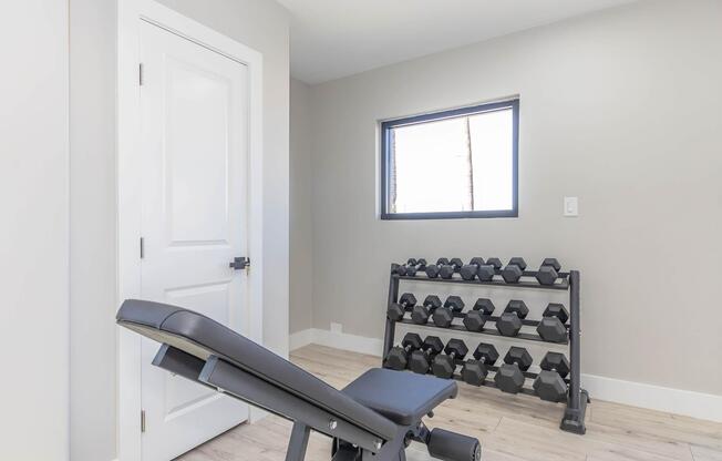A minimalistic home gym featuring a weight bench and a black dumbbell rack filled with various weights. A window lets in natural light, and a white door is visible on the left, adding to the modern and clean aesthetic of the space.