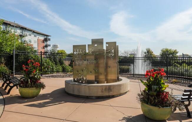 A fountain in the middle of a park with benches and flower pots.