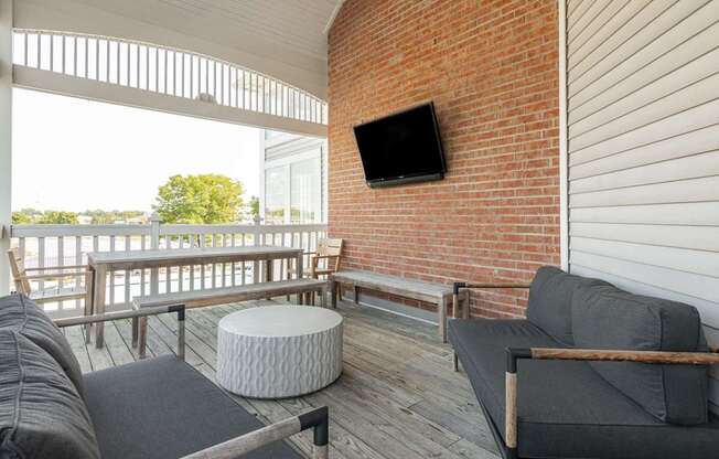 A balcony with a grey couch, a white ottoman and a flat screen TV mounted on a brick wall.