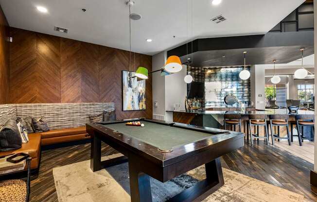 A pool table is in the middle of a room with a wooden floor and a bar area.