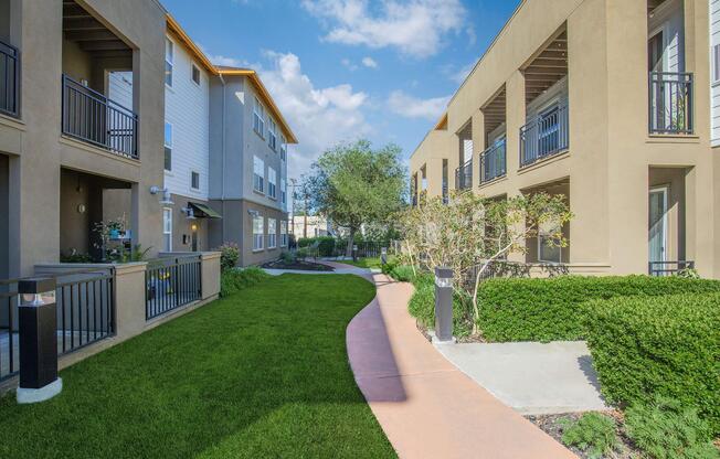 A walkway lined with green grass and neatly trimmed bushes, leading through a modern apartment complex with two buildings. The sky is partly cloudy, and the architecture features balconies and light-colored facades.
