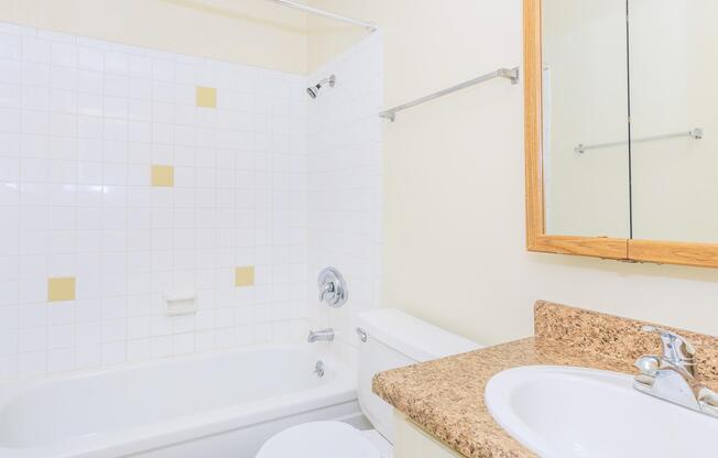 A clean, well-lit bathroom featuring a bathtub with a shower, white tiled walls with yellow accents, a beige countertop with a sink, and a mirror above. The space is tidy and minimalistic, emphasizing a neutral color palette.
