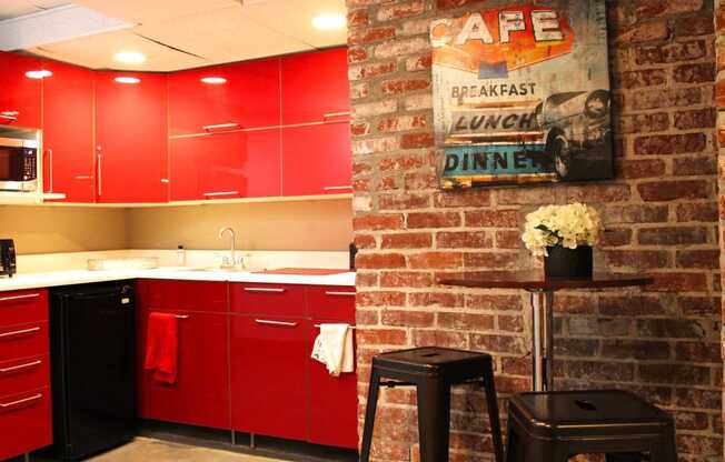 Brookmore apartments community kitchen with red cabinets, dishwasher, and microwave, and sitting area with small bar table and two black stools.