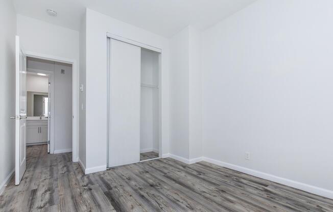 A bright, empty room with white walls and a light wood floor. There is a mirrored closet door on the left. A doorway leads to another room, partially visible, which appears to include a bathroom area. The overall atmosphere is clean and minimalistic.