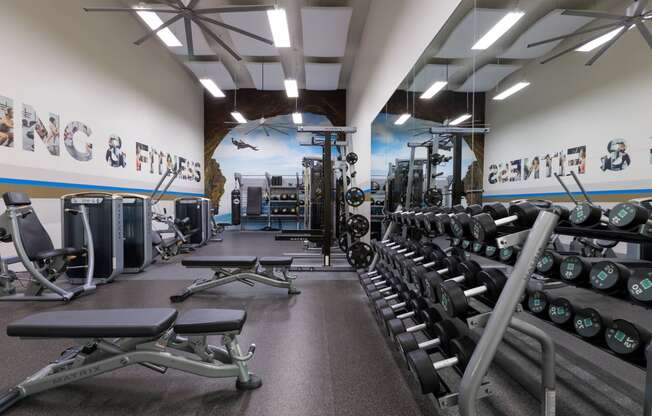 a gym with cardio machines and weights on the floor and a wall with a mural