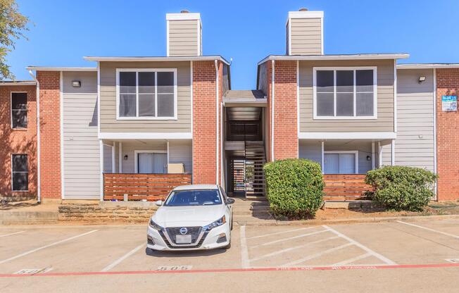 Two-story apartment building with a brick facade and light-colored siding. The entrance has large windows, and there is a white car parked in front. Green bushes flank the sides of the building, and parking spaces are marked in front. The scene is bright and sunny.