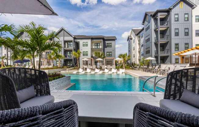 A pool surrounded by chairs and umbrellas in front of apartment buildings.