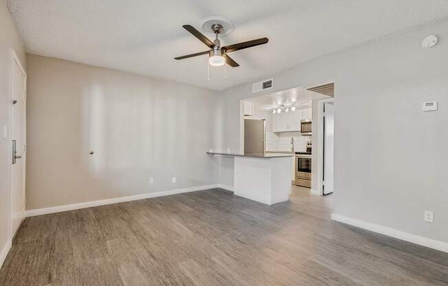 A spacious room with a ceiling fan and light fixture, and a kitchen area visible through the open door.