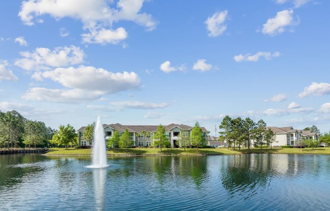 A fountain in the middle of a lake with houses in the background.