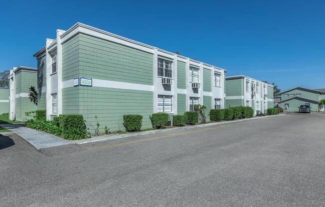 A residential building with a green exterior, featuring multiple units with windows, shrubs along the walkway, and a clear blue sky. The area includes a paved road and another structure in the background.