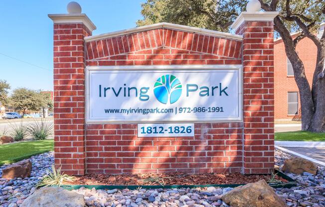 Brick sign for Irving Park, featuring the logo and address "1812-1826" along with the website and phone number. Surrounding the sign are decorative stones and landscaping elements.