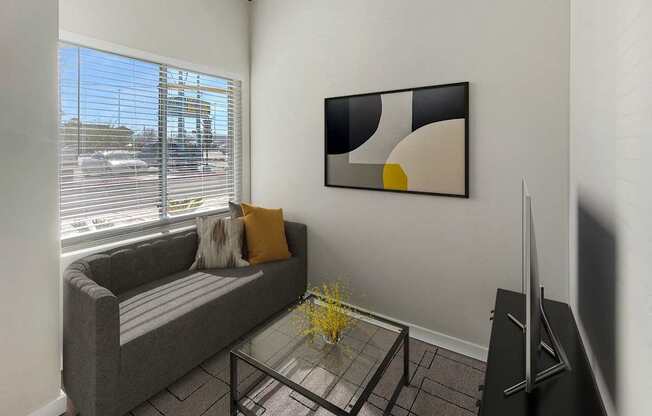 A living room with a grey couch, a glass coffee table, and a painting on the wall.