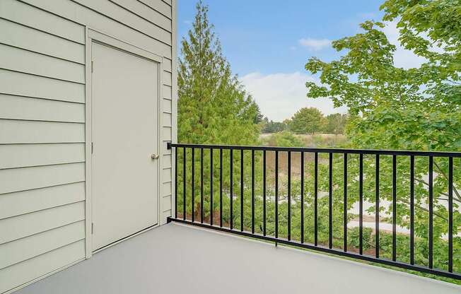 A private outdoor balcony here at Heritage Hills featuring a smooth patio floor, black metal railing, and light exterior siding with a storage door. The balcony overlooks mature trees and landscaped greenery, offering a peaceful outdoor space with natural views and fresh air just outside the apartment home.