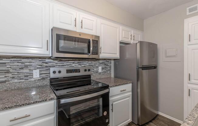 A modern kitchen featuring white cabinetry, granite countertops, a stainless steel refrigerator, an oven with a glass top, and a microwave above the stove. The backsplash consists of decorative tiles in various shades, providing a stylish contrast to the overall design.